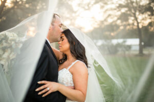 A bride in a lace wedding dress and a groom in a black tuxedo suit holding each other.