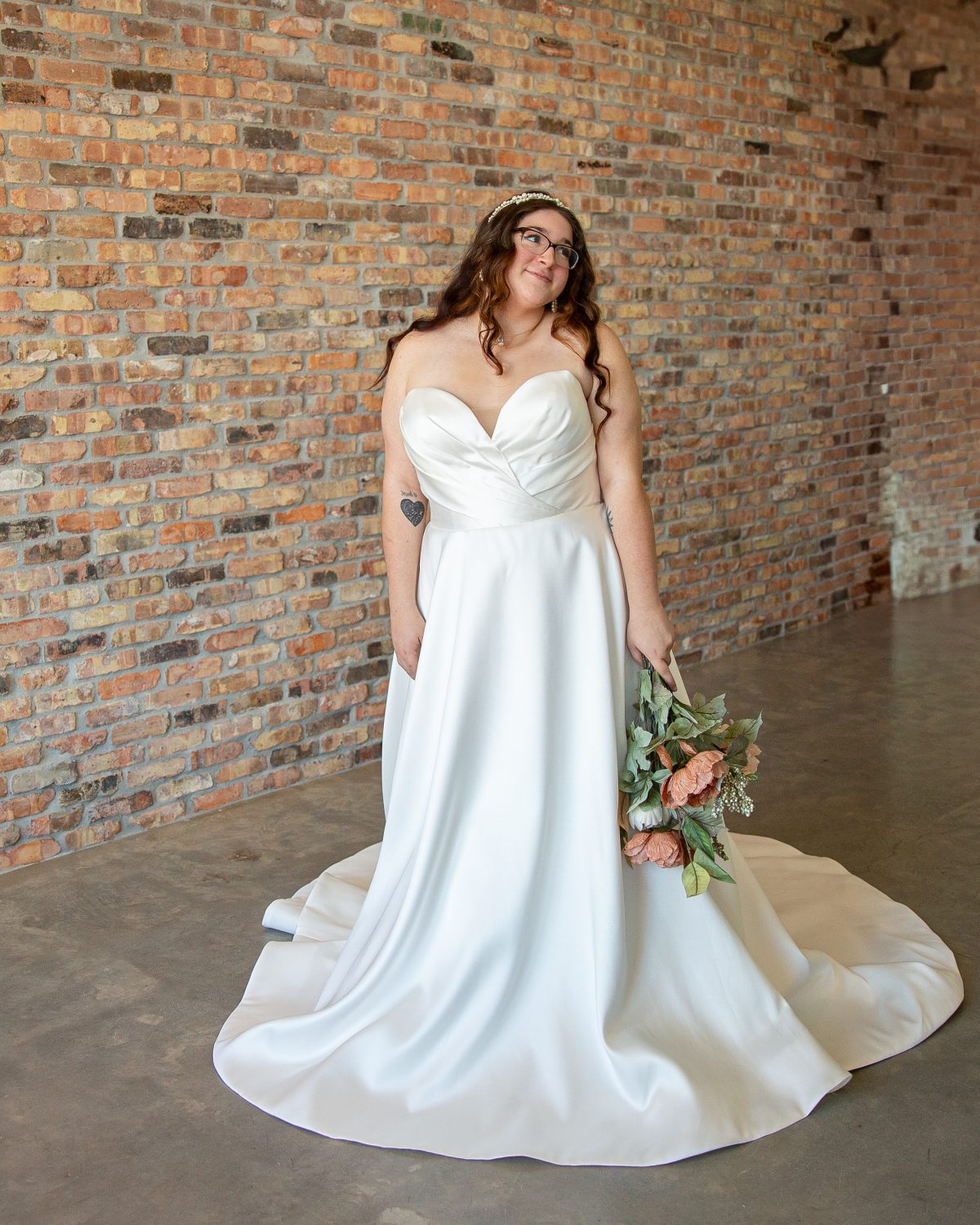 Plus-size bride wearing the Grace wedding dress in ivory, featuring an A-line silhouette, strapless sweetheart neckline, ruched bodice, pockets, and detachable sleeves. Bride poses modestly in a bashful, shy stance. Timeless Collection at Satin and Lace Bridal Boutique in Riverview, FL.