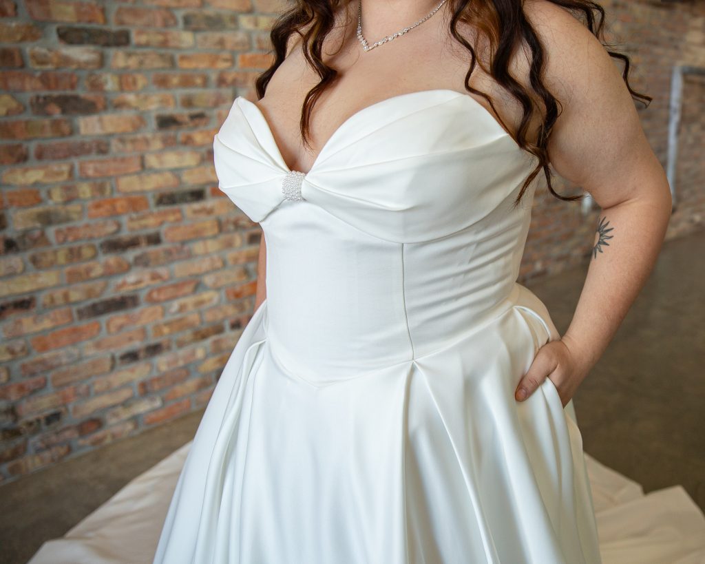 Close-up of plus-size bride wearing Grace wedding dress from Timeless Collection at Satin and Lace Bridal Boutique, Tampa, Riverview, FL. Ivory A-line dress with drop waist, strapless bow neckline, detachable straps, pearl detailing, and pockets.