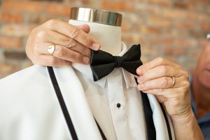 Bridal boutique owner adjusting a black bowtie on a mannequin wearing a classic white tuxedo with black trim, customizable suit rental option in Tampa, Florida.