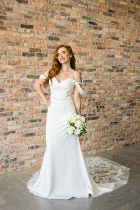 Front view of a long haired bride wearing Merida wedding dress in Off white with off the shoulder straps, hand on hip, holding a floral bouquet in front of a brick wall at Satin and Lace Bridal Boutique in Riverview Florida