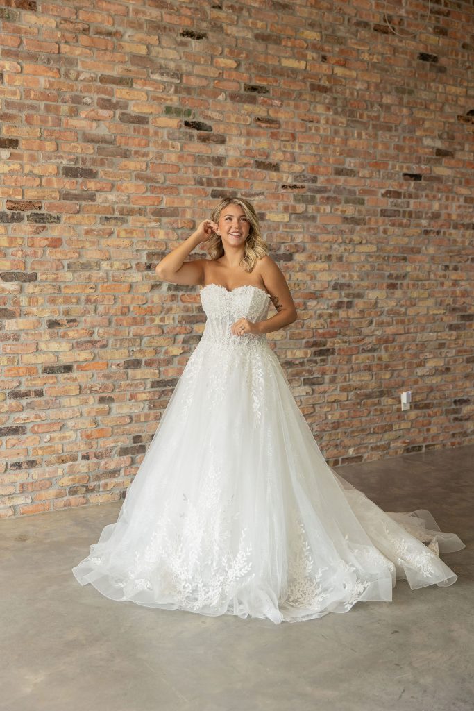 Front view of bride with blonde hair pushing her hair behind her ear, wearing Annabelle an A line strapless lace and sparkle wedding dress without the detachable cuff sleeves, in front of a brick wall at Satin and Lace Bridal Boutique Tampa Florida