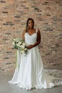 Front view of a smiling dark skinned bride with long braids wearing an A line satin gown with sequined and beaded appliques on the bodice, a lace cutout train with scalloped edge and 3D lace straps holding a bouquet of flowers in her right hand, in front of a brick wall at Satin and Lace Bridal Boutique Tampa Florida.