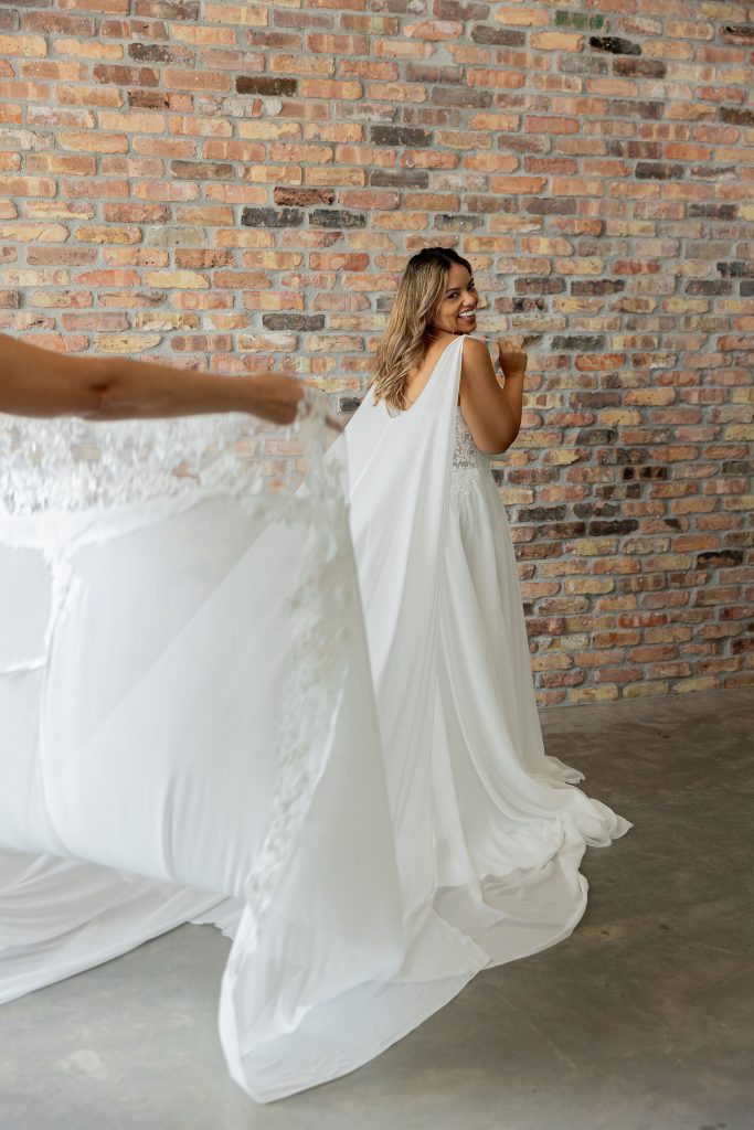 Shows back view of a smiling bride looking over her shoulder wearing a 2 piece A Line dress with Basque waist. Out of shot an assistant is holding the end of the cape to showcase the flowing cape, in front of a brick wall at Satin and Lace Bridal Boutique Riverview Florida