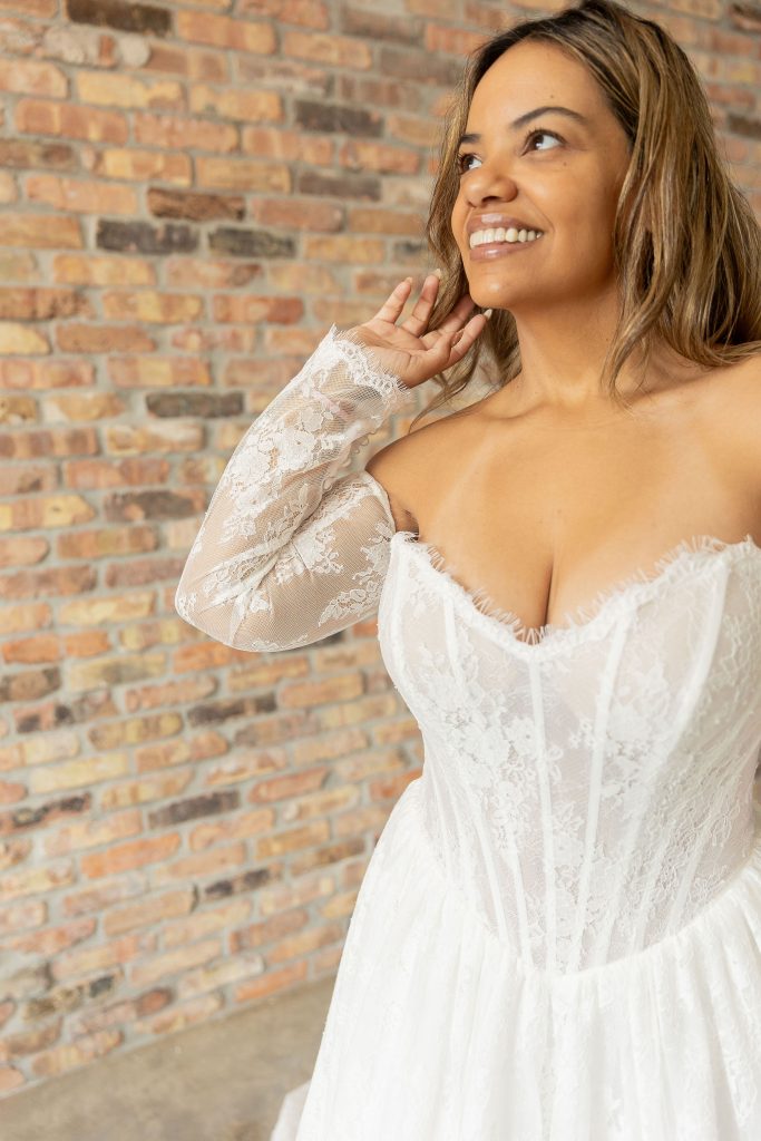 Close up Front view of a smiling Hispanic bride with long hair, wearing Nadia an A line Basque waist strapless Chantilly lace wedding dress with eyelash lace across the top of the bodice and separate long lace sleeves, in front of a brick wall at Satin and Lace Bridal Boutique Tampa Florida